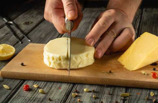istock Hands elegantly cut a round cheese on a wooden board, surrounded by spices and lemon. The warm ambiance highlights the artisanal process of cheese preparation, evoking culinary creativity 2199855401