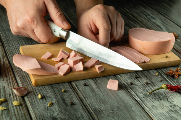 istock An experienced hand skillfully cuts a piece of cooked sausage with a sharp knife, preparing small cubes on a wooden cutting board 2199855488
