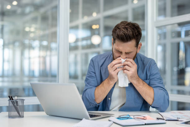 istock Sick man suffering at workplace, sneezing. Worker has runny nose and cold blowing snot into a napkin. Working with laptop inside office 2199859771