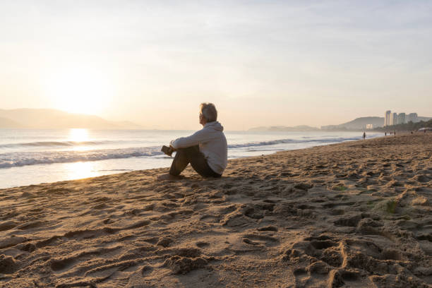 Mature man watches sunrise from beach holding mobile phone, islands and mountains distant, Nha Trang