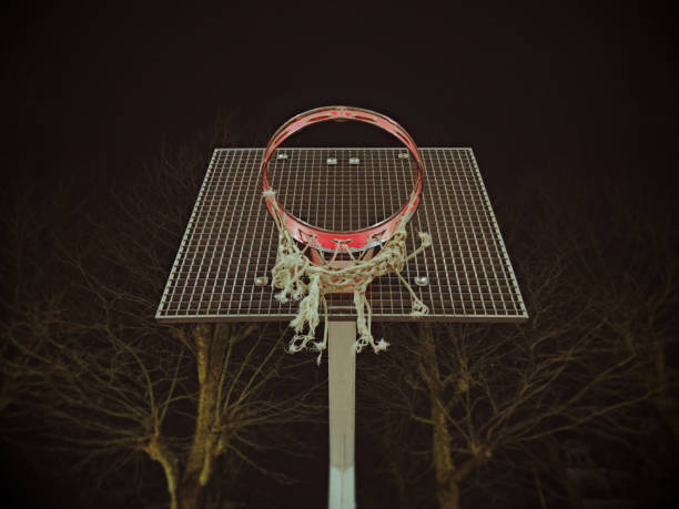 a basketball hoop attached to a metal mesh backboard, mounted on a vertical support. The hoop has a red rim, and the net is significantly frayed with several loose strands. Bare tree branches are visible in the dark background, indicating a nighttime setting.