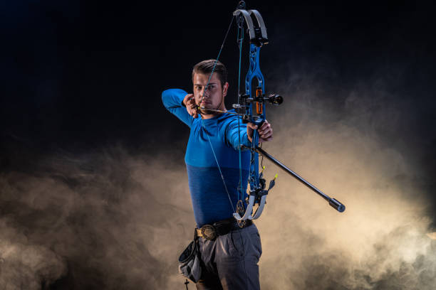 Young Caucasian male archer in a studio setting aiming an arrow with precision. Dressed in a blue shirt, he demonstrates skill and concentration. The backdrop is a dramatic blend of dark and light, enhancing the intensity.