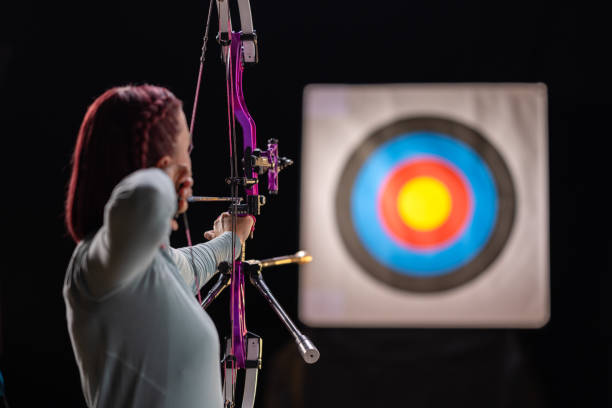 A mid adult Caucasian female archer aims her bow in a studio environment. She wears a light blue top and holds a modern purple bow. The target in the background is sharply defined against the dark backdrop.