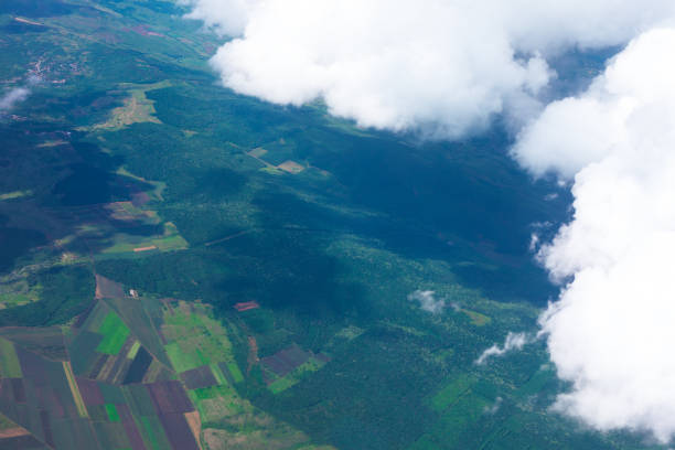 Aerial view of a lush green landscape with agricultural fields and dense forests, partially covered by fluffy white clouds casting shadows below. Mix of cultivated land and vibrant vegetation