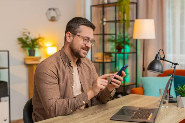 istock Happy young male freelancer sitting at desk using smartphone texting smiling at home apartment 2199889401