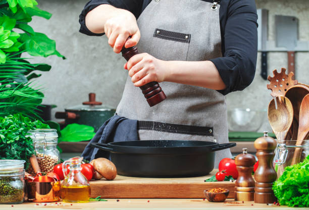istock Chef grinds salt in spice grinder over cast iron skillet. Cozy kitchen with wooden table, kitchenware, vegetables, herbs and ingredients for cooking. Healthy food, eating, culinary, recipes, food blogging 2199919238