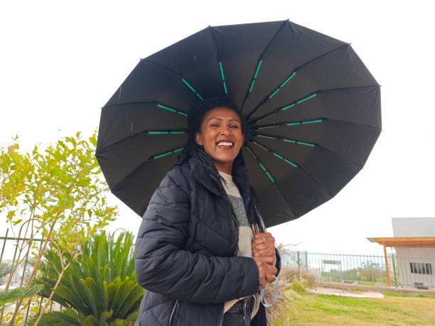 A stylish woman with a warm smile poses outdoors while holding a black umbrella. Dressed in a cozy black jacket and a striped sweater, she enjoys the fresh air despite the rainy weather. The background features lush green plants and a modern urban environment, adding a contemporary feel to the image. The umbrella frames her face, creating a visually appealing composition that captures confidence and joy in an outdoor setting.