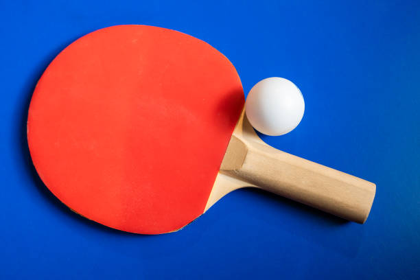 Looking down on four multi-colored plastic ping-pong paddles and an orange ball on a blue table tennis surface.