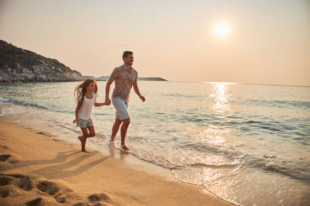 istock Father and daughter run hand in hand down a tropical paradise beach during sunset 2199935075
