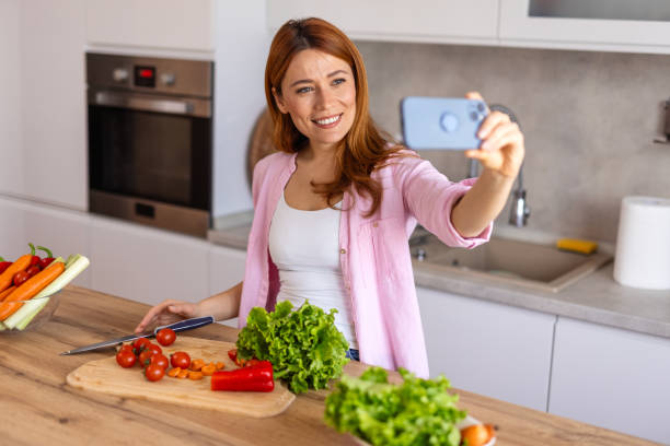 istock A woman in a modern kitchen prepares dinner while taking a selfie, capturing a fun and joyful moment. She is multitasking, cooking a delicious homemade meal while enjoying the process. 2199939046