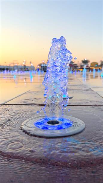 Fountain jets rise from ground, illuminated by blue lights, shimmering droplets reflecting evening glow, modern illuminated fountain display creates dynamic patterns, urban ambiance concept.
