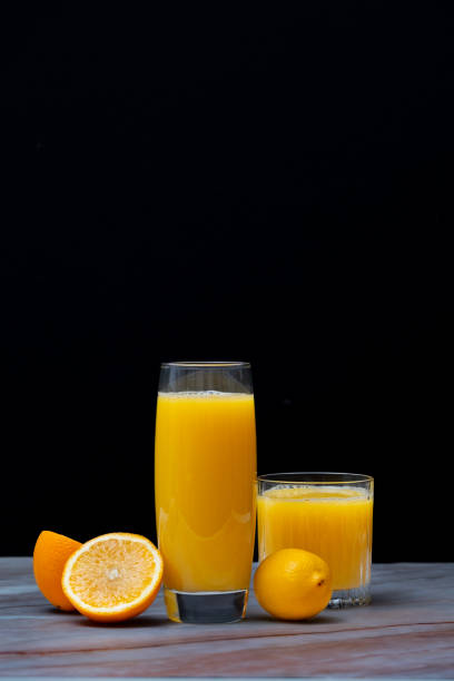 Two glasses of orange and lemon juice on a pink marble table with two halved oranges and a lemon, black background, front view, vertical image.