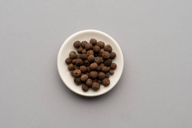 A top-down view of aromatic spices and dried tea leaves arranged on a white background. The composition features star anise, black peppercorns, and two small ceramic bowls filled with dried tea leaves, creating a minimalist yet elegant food presentation. The scattered elements add an organic feel, emphasizing the rich textures and natural beauty of these culinary ingredients. This image is ideal for use in food blogs, tea packaging designs, spice advertisements, or educational content about traditional herbs and spices.
