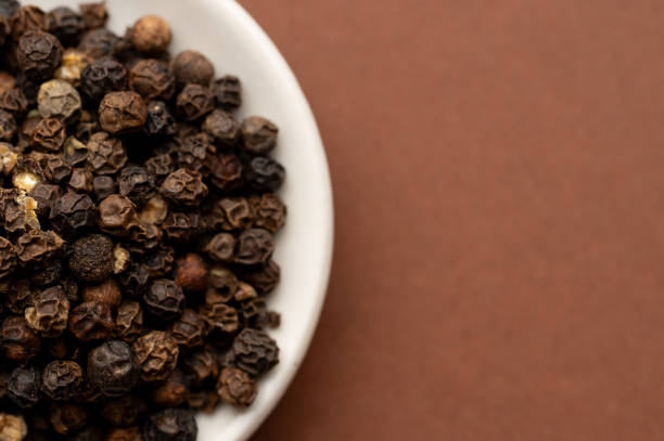 Top-down view of black peppercorns in a white ceramic bowl on a brown background. The round peppercorns and smooth bowl create a simple, minimalistic culinary composition highlighting natural spices.