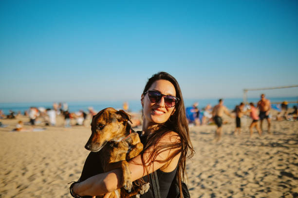 Woman in her late 30s, early 40s, relaxing at the beach in Barcelona in the afternoon, under the beautiful sunlight, enjoying the staycation with her small pet dog. Pet friendly beaches, staycations and city break concepts in Europe.