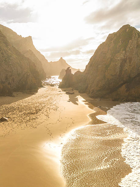 Dramatic coastal landscape featuring towering rock formations, golden sand, and ocean waves crashing against the shore under a warm sunset. Stock photo