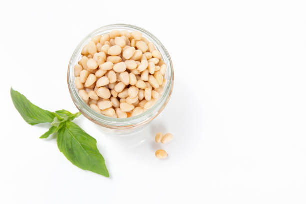 A jar of pine nuts with fresh basil leaves on white background, great for homemade pesto, salads, and Mediterranean dishes
