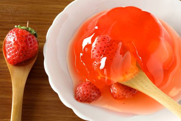 Close-up of fresh strawberries in red jelly on a white plate with a wooden spoon on a wooden table.