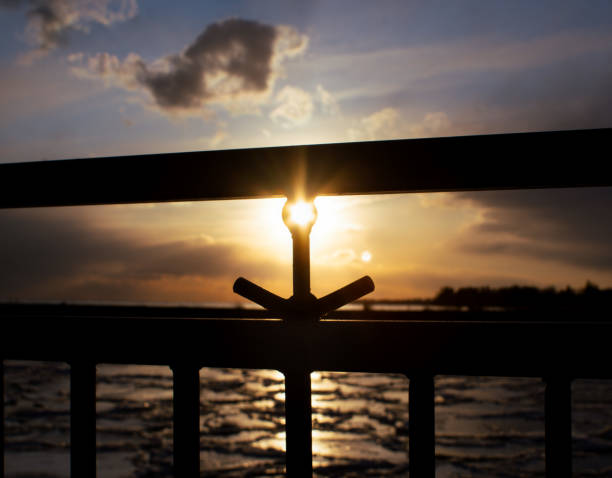 The sun sets behind a metal railing, perfectly aligning with a small gap, casting a golden glow over a rippling body of water. A serene moment blending nature and urban structure.