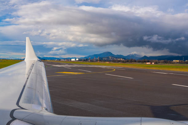 An airplane wing stretched over a runway, highlighting the expansive green landscape and distant mountains under a cloudy sky.
