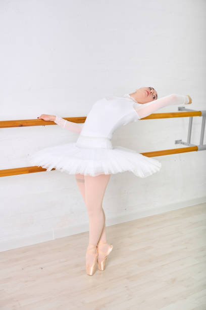 Young female ballerina with short hair gracefully stretches at barre in dance studio. Clad in white tutu, poised with elegance under soft lighting and white background
