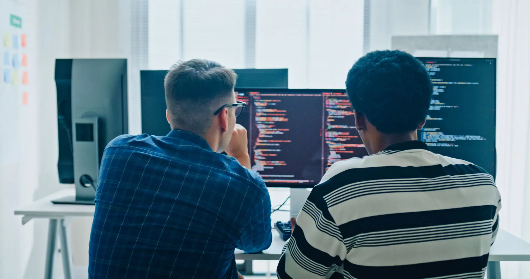 Two male software developers meeting on multiple computer screens displaying programming code, collaborating on project in office. Artificial intelligence and programming.