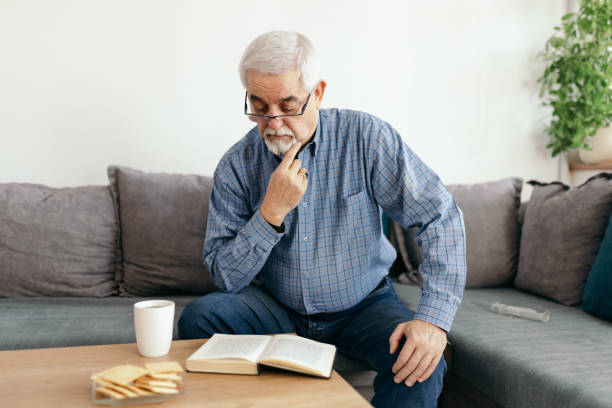 Senior man spending his pension time as best as he can. He is using free time to read a book in a living room at home.