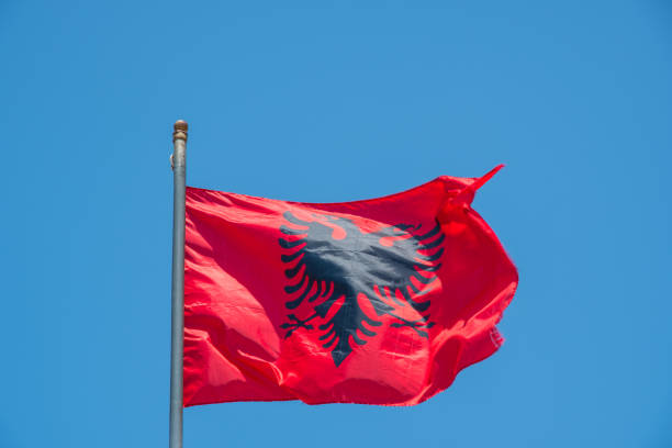 Albanian national flag waving on a tall pole against blue sky with clouds and mountains at sunset