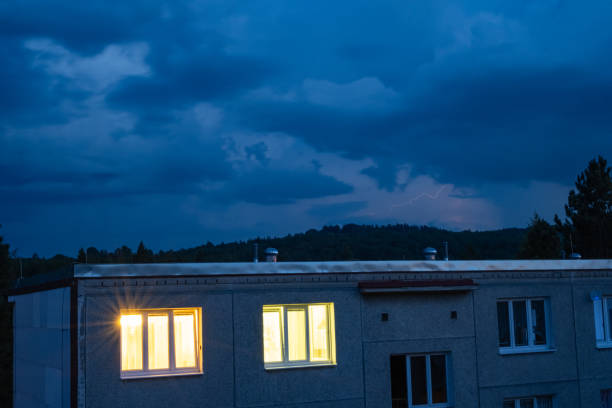 A dramatic evening sky with dark storm clouds and a faint lightning strike in the distance. The foreground features a residential building with illuminated windows, contrasting with the moody atmosphere.