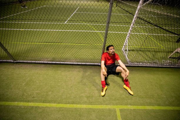 solitary soccer player sits on the grass, gazing down in exhaustion, next to a chain fence after a loss.