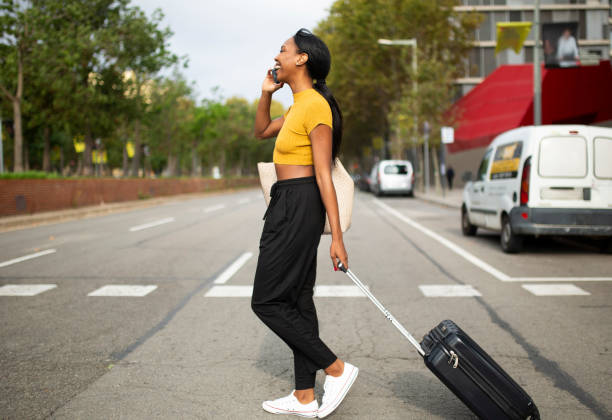 istock Black woman talking on mobile phone while pulling luggage and crossing road 2200065354