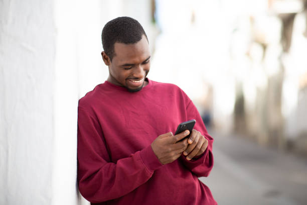 istock Smiling black guy leaning to wall and texting on mobile phone outside 2200065748