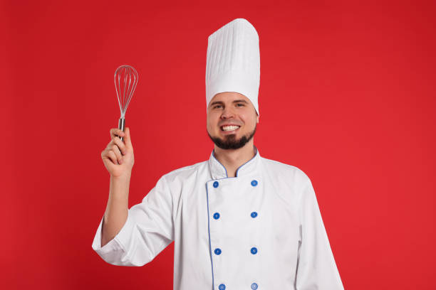 istock Happy professional confectioner in uniform holding whisk on red background 2200085841