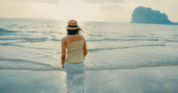 istock Young Asian woman walking barefoot along on sandy beach during sunset, enjoying tranquil waves and warm sunlight, evoking relaxation. Vacation destinations and Lifestyle tropical beach. 2200091907