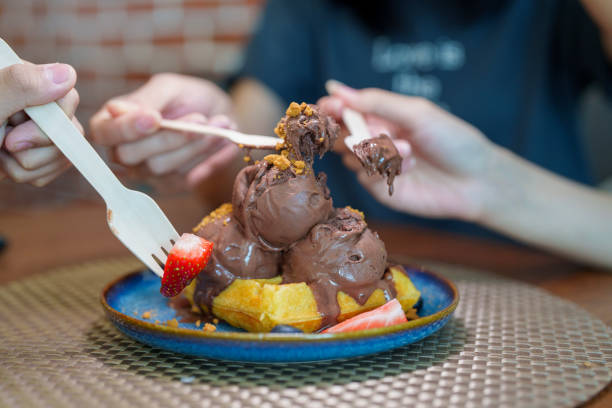 Close-up of chocolate ice cream scoops on crispy waffles, garnished with strawberries and biscuit crumbs, shared among friends in a cafe.