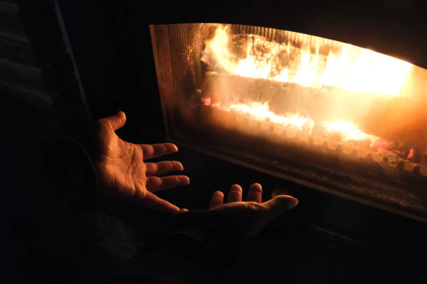 Two hands are held up close to a glass-enclosed fireplace, with flames visible inside, suggesting warmth and comfort. The scene is cozy and inviting.