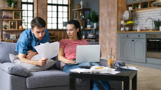 istock Couple calculating bills at home using tablet and calculator. Young couple working on computer while calculating finances sitting on couch. Young man with wife at home analyzing their finance with documents 2200116408