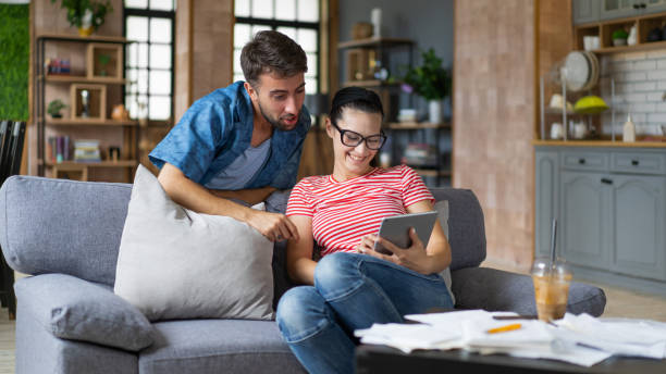 istock Couple calculating bills at home using tablet and calculator. Young couple working on computer while calculating finances sitting on couch. Young man with wife at home analyzing their finance with documents 2200116410