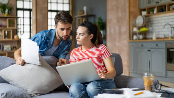 istock Couple calculating bills at home using tablet and calculator. Young couple working on computer while calculating finances sitting on couch. Young man with wife at home analyzing their finance with documents 2200116416