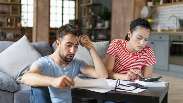 Couple calculating bills at home using tablet and calculator. Young couple working on computer while calculating finances sitting on couch. Young man with wife at home analyzing their finance with documents
