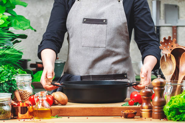 istock Housewife holds cast iron saucepan in hands. Cozy kitchen with wooden table, kitchenware, vegetables, herbs and ingredients for cooking. Healthy eating, culinary, recipes, food blogging 2200121103