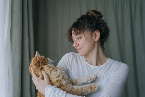 istock Woman holding ginger cat near window with curtains 2200126571