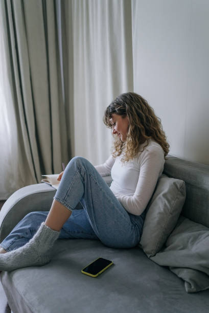istock Young woman writing in notebook sitting on sofa at home 2200127643