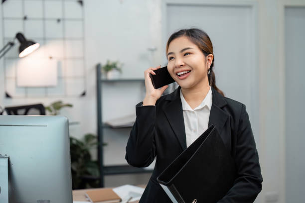 istock Enthusiastic Asian female call center agent talking on the phone while holding a folder in a modern workspace, showcasing engagement. 2200127772