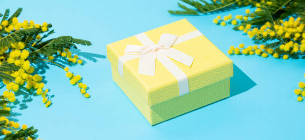 istock Yellow gift box on blue background with mimosa flowers and sunlight. 2200132728