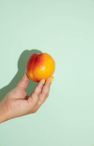 istock Vertical image of female hand holding a peach on a light green background with copy space 2200133242