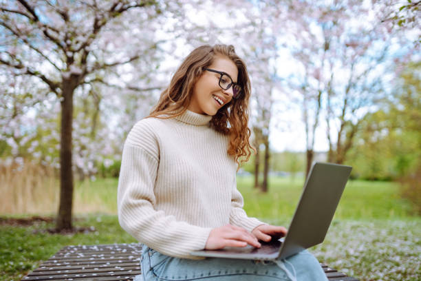 istock Young woman sitting on blooming park and using laptop. 2200134849