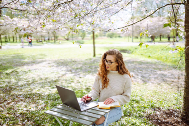 istock Young woman sitting on blooming park and using laptop. 2200134862