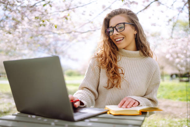 istock Young woman sitting on blooming park and using laptop. 2200134863