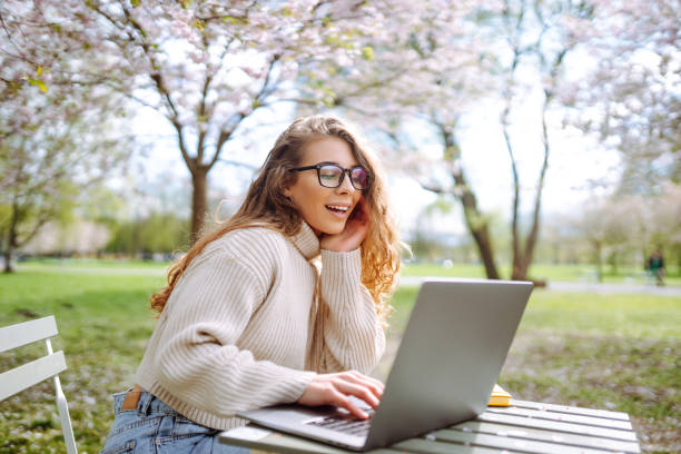 istock Young woman sitting on blooming park and using laptop. 2200134869